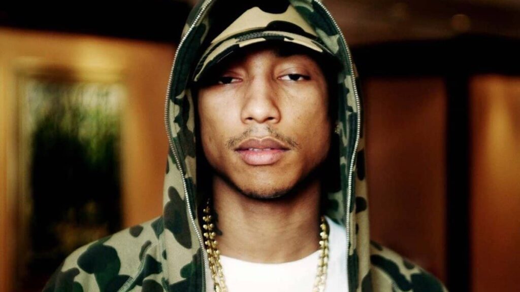 Pharrell Williams biography portrait — posing in a stylish hat and jacket.