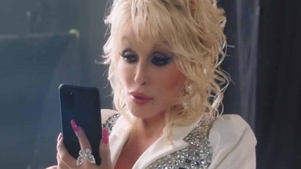Dolly Parton biography portrait — smiling in a pink dress and holding a phone during a 2022 commercial shoot.