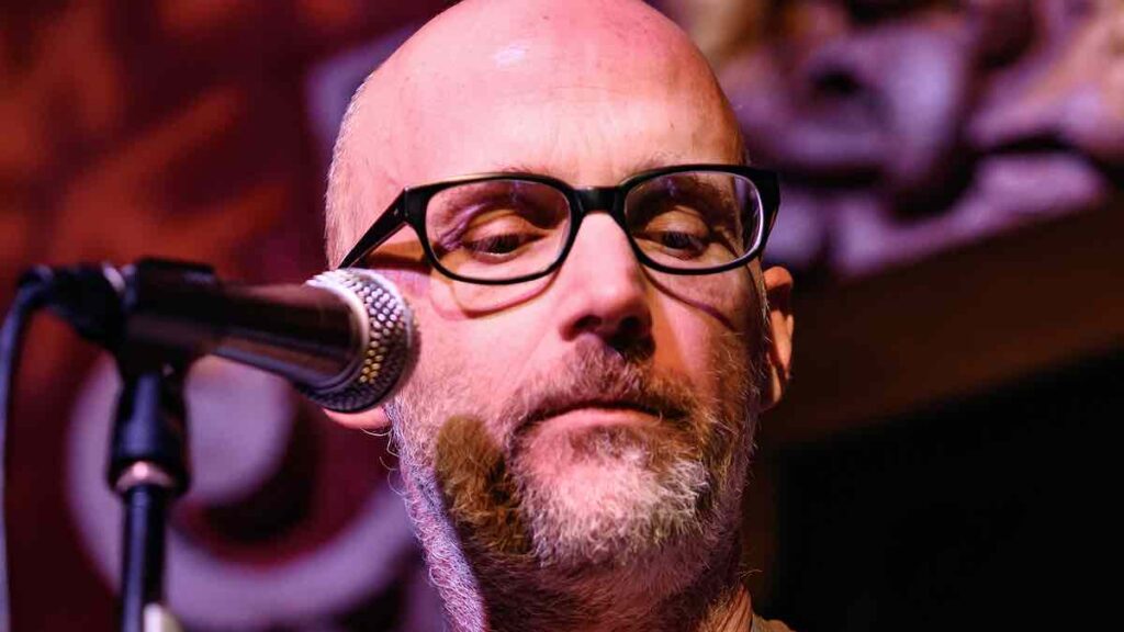 Moby biography portrait — performing live with a guitar on stage during a concert.