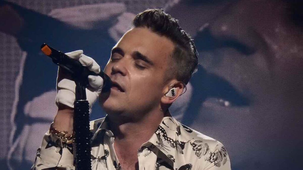 Robbie Williams biography portrait — singing passionately into a microphone during a concert in London.